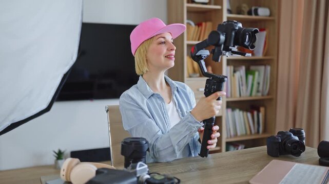 A young woman with blonde hair and a pink cap is setting up her camera on a gimbal stabilizer in a home studio environment