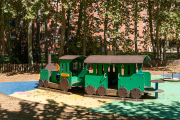 A children's playground features a green wooden train with a locomotive and connected carriage designed for imaginative play. Colorful safety flooring, surrounding shade trees and residential building © Volha