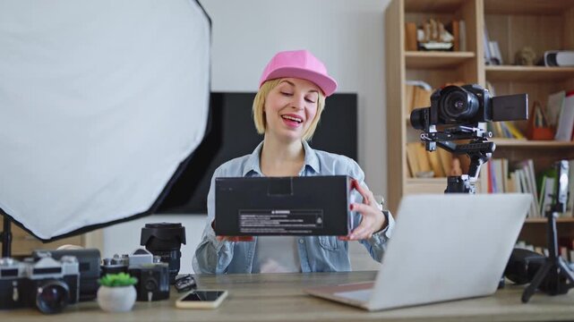 A smiling caucasian woman wearing a pink cap presents a black box to the camera in her home studio setup with professional equipment