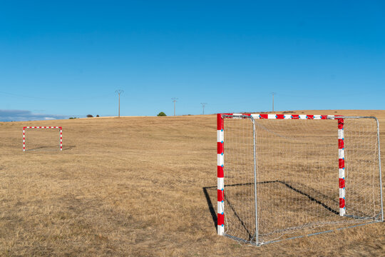 Two small football goals stand far apart on a sun-dried grass field surrounded by open countryside. Their red and white metal frames and netting contrast with the golden vegetation, creating a scene o