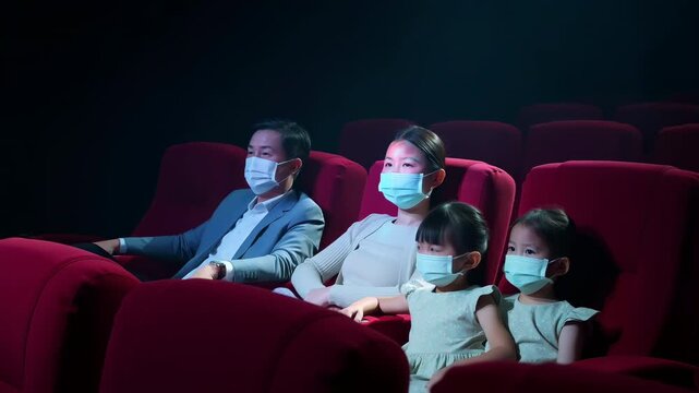 Audience members wearing face masks sit in rows of red theater seats while watching screen in dark cinema interior with cool blue lighting, quiet viewing atmosphere, and side perspective composition