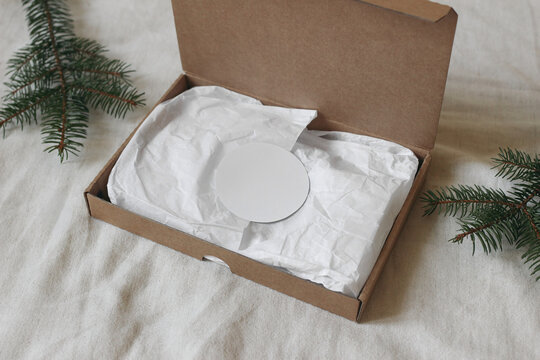 Christmas Gift Packaging Mockup with Blank Round White Sticker Label on Tissue Paper in Open Kraft Cardboard Box. Fir Branches on Natural Linen Blurred Background, Eco Friendly Holiday Branding