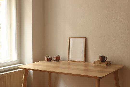 Minimalist Autumn Home Interior. Blank Wooden Picture Frame Poster Mockup on table near window. Rustic Pumpkins. Ceramic Coffee Cup, Books. Scandinavian Fall Decor, Wooden Table by Window, Beige Wall