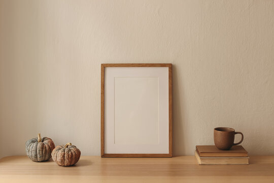 Minimalist Autumn Home Decor with Blank Wooden Picture Frame Poster Mockup. Two Pumpkins and Brown Ceramic Coffee Cup on Vintage Books. Cozy Scandinavian Fall Interior, Neutral Beige Wall Background