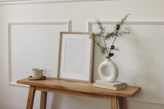Minimalist Scandinavian Home Interior. Blank Wooden Picture Frame Poster Mockup. Modern Ceramic Donut Vase with Larch Branches Pine cones. Rustic Wooden Bench Books Coffee Cup. Nordic Christmas Decor