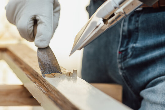 Adult carpenter wearing protective leather gloves, using a heat gun and a wood scraper to remove paint from an old wooden window. Construction industry, carpentry. Restoration. 