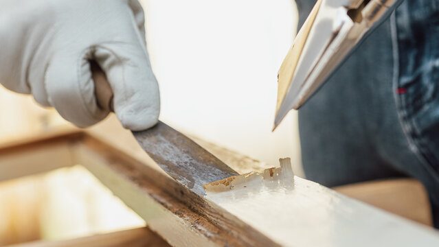Adult carpenter wearing protective leather gloves, using a heat gun and a wood scraper to remove paint from an old wooden window. Construction industry, carpentry. Restoration. 