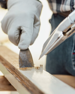Adult carpenter wearing protective leather gloves, using a heat gun and a wood scraper to remove paint from an old wooden window. Construction industry, carpentry. Restoration. 