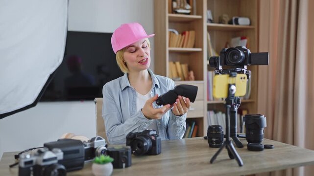 A caucasian woman in a pink cap explains camera equipment while recording a video with a smartphone and DSLR camera setup