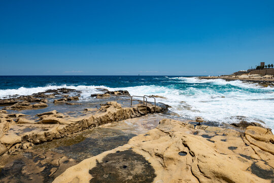 White sea foam and splashing waves crashing over natural tide pools with distant watchtower in Sliema Malta