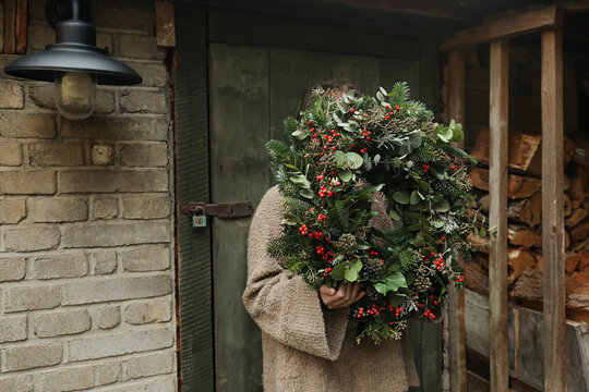 Child Holding Large Handmade Christmas Wreath with Fir Branches, Eucalyptus and Red Berries Outside Rustic Country Cottage with Old Brick Wall, Wooden Door and Firewood, Natural Holiday Decoration