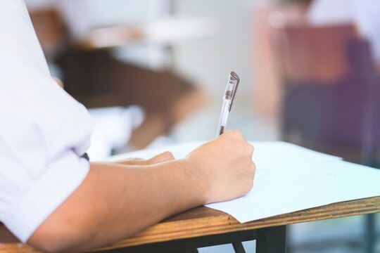 Close-up of a student's hand diligently writing on an exam paper during a school test. 
The student is using a professional pen , sitting at a wooden desk in a brightly lit classroom.
