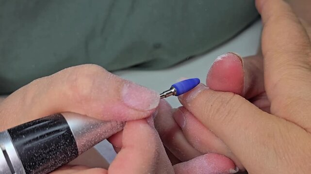 Close up of a manicurist using an electric nail drill with a blue ceramic bit to remove old gel polish from a woman's nail in a beauty salon, grainy film texture