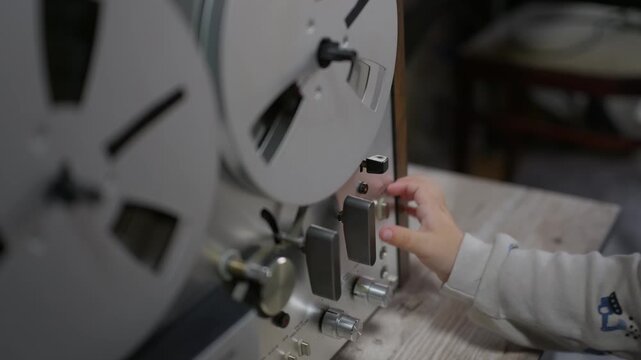 A curious child operates a vintage reel-to-reel tape recorder indoors in a close-up. Little hands carefully toggle switches and press control buttons on the retro analog audio deck.