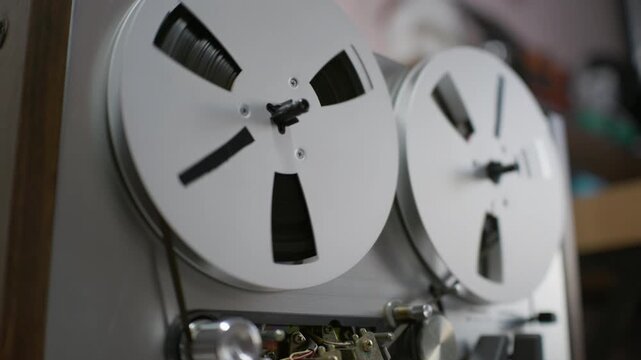A vintage reel to reel tape recorder spins magnetic audio tape reels inside a recording studio. Spools turn smoothly as mechanical components operate under soft ambient indoor lighting.