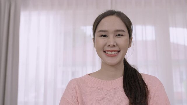 Happy, confident, beautiful young Asian woman in pink clothes standing smiling and looking at the camera in the living room at home.  Portrait of a pretty Thai female model in a studio. 