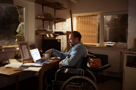 Smiling man in wheelchair works on laptop at home with dog