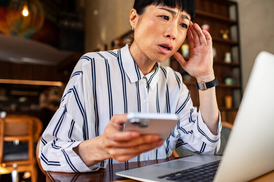 Worried woman holding phone while looking at laptop in cafe
