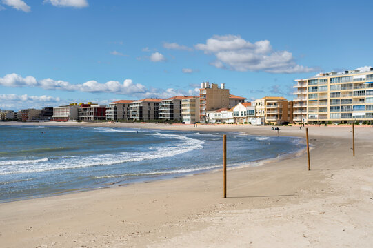 A wide sandy beach stretches along the Mediterranean coastline in Palavas-les-Flots, France. Modern apartment buildings and seaside hotels front the water under a bright sky.
