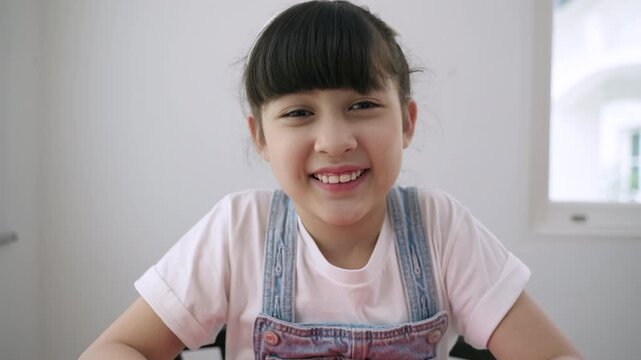 Cute Asian girl student sitting at desk is looking at the webcam while talking to computer camera and video calling with a remote teacher for online learning at home. Homeschooling education concept. 