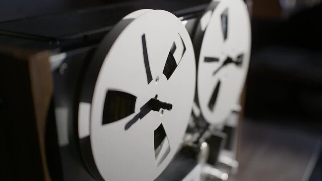 Metal spools on a vintage reel-to-reel tape recorder spin steadily during analog audio playback. Strong side illumination casts deep cinematic shadows across the revolving metal reels.