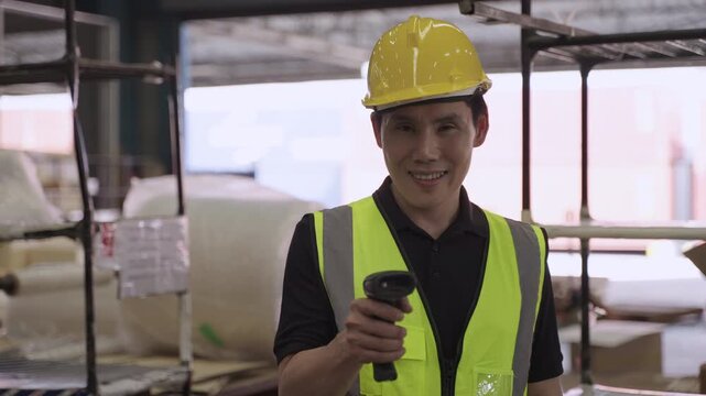 Portrait of happy Asian man warehouse worker in uniform and wearing yellow safety helmet, standing and holding barcode scanner while looking at camera in a logistics distribution factory. 