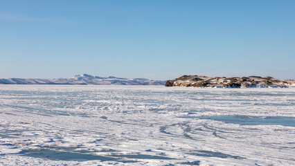 Vast icy expanse of frozen Lake Baikal covered with snow patterns under a clear blue winter sky in Irkutsk Oblast, Russia, showing rocky hills and mountain ranges along the Siberian horizon © TravelFlow