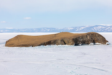 Rocky Kharantsy Island stands in the vast expanse of frozen Lake Baikal in Irkutsk Oblast, Russia, surrounded by ice and snowy distant mountain ridges under a bright winter sky © TravelFlow