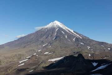 Majestic stratovolcano with snow covered slopes rises above rugged volcanic terrain under clear sky in Kamchatka Krai, Russia, showcasing pristine wilderness and dramatic natural landscape © TravelFlow