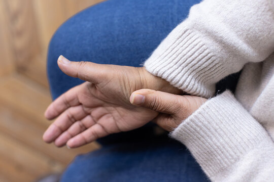Mature African American woman resting hands on lap, wearing cream sweater and jeans, grasping wrist