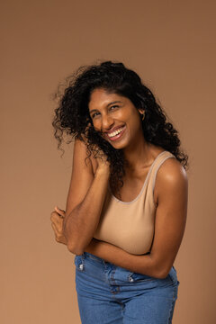 Woman smiling, touching hair, posing in studio with ribbed tank, high-waisted jeans, tan backdrop