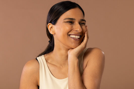 Indian woman laughing, resting hand on cheek, wearing sleeveless beige top against beige backdrop