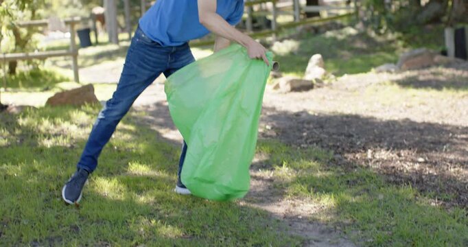 Diverse male friends in blue tees spotting tube and bottle, scooping into green bag cleaning park