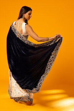Adult Indian woman posing, lifting navy velvet pallu with gold border in studio on orange backdrop