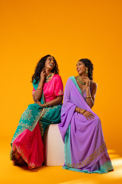 Diverse female friends sitting on white stool against orange backdrop wearing saris and jewelry