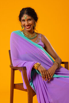 Indian woman sitting and smiling on wooden chair in studio, wearing purple saree, gold jewelry