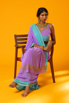 Indian woman wearing lavender sari with maang tikka, bangles, sitting on wooden chair in studio