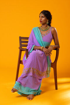 Indian woman sitting on wooden chair in studio wearing lavender turquoise outfit and gold jewelry