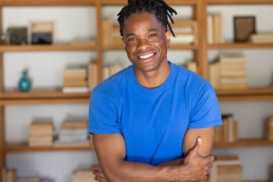 Man smiling, standing with arms crossed at home by wooden shelving with books wearing blue T-shirt