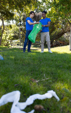 Diverse male volunteers wearing blue shirts collecting litter in park with green trash bag