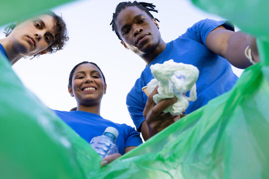 Volunteers leaning over green trash bag at park, wearing blue T-shirts holding bottle