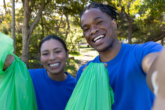 African American volunteers wearing blue shirts holding green trash bags and taking selfie in park
