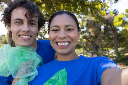 Diverse volunteers taking phone selfie in park wearing blue tees holding translucent green bags