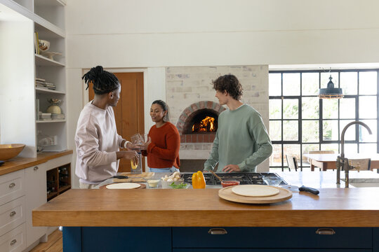Diverse friends handing pitcher and preparing dough on wood island in home kitchen with bell pepper