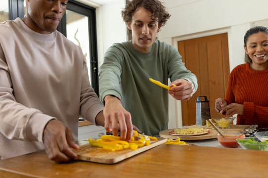 Diverse friends chopping yellow peppers on board and topping pizzas on peel at kitchen island