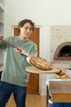 Male cook in green sweatshirt passing pizza with metal peel to recipient by brick oven