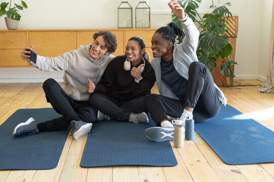 Friends posing for selfie in sportswear on yoga mats at home, holding smartphone and water bottles