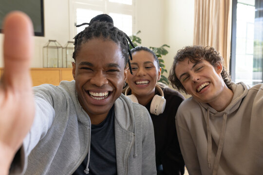 Diverse friends taking selfie on couch in living room with smartphone and white headphones visible