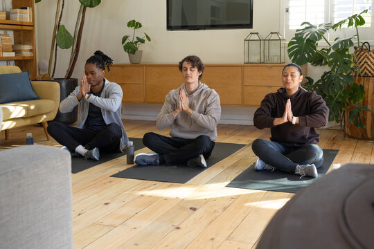 Diverse friends meditating on black yoga mats in living room, wearing hoodies, water bottles nearby