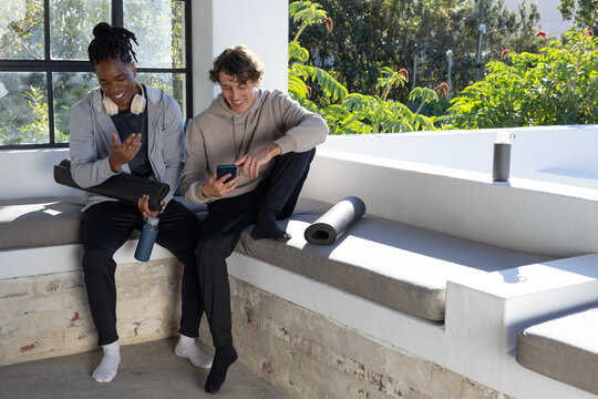 Diverse male friends sitting in alcove in athleisure, sharing smartphone, headphones, water bottle